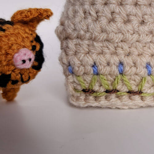A close up of a crocheted spotted piglet next to the embroidery on the crocheted sty