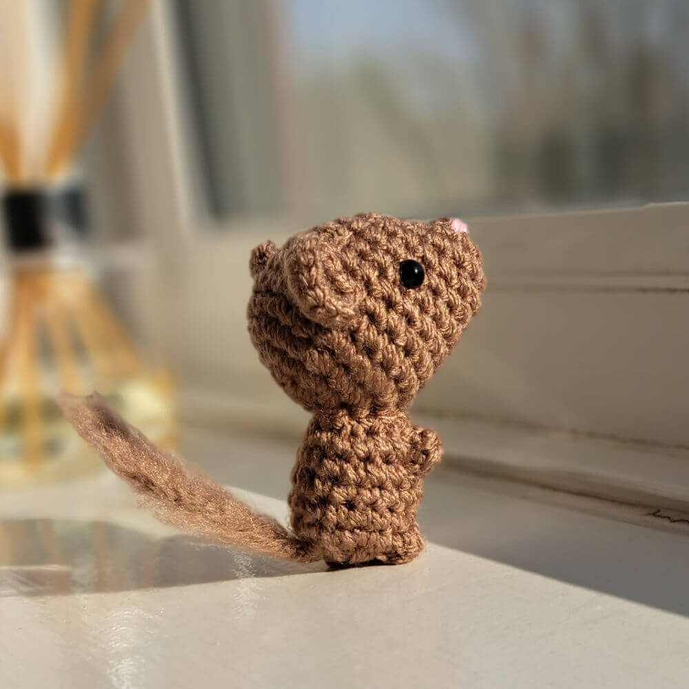 A super cute little brown mouse crochet figure with a fluffy tail is looking out of a window on a sunny day.