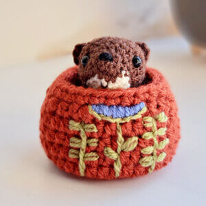 An adorable little brown weasel ornament crochet art figure peeping out of his cosy embroidered nest on a window sill