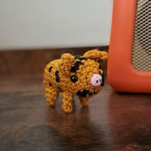 An adorable little spotted piglet crochet figure standing on a shelf with a radio in the background
