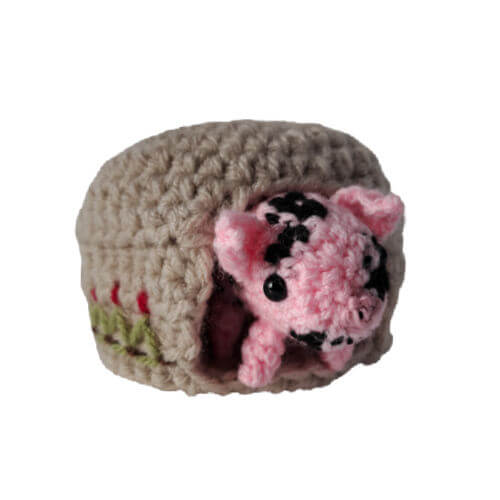 A cute little crochet pink piglet with black spots peeping out of her embroidered sty