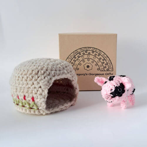 A pink piglet with black spots standing with an embroidered sty and gift box