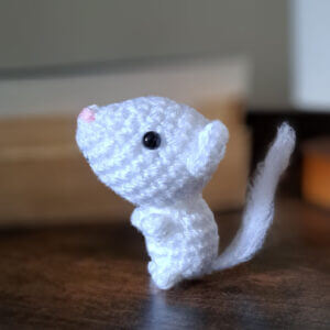 A super cute white mouse ornament crochet figure sitting up on a bedside table with books in the background.