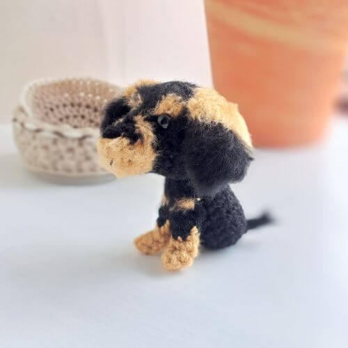 A wire-haried dachshund crochet art figure sitting on a window sill looking into the room, there is a crochet basket and a plant pot in the background