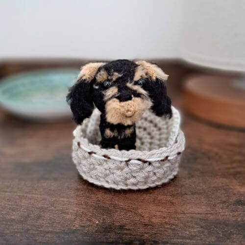 An adorable little wire-haired dachshund crochet art figure sitting in his little crochet basket on a bedside table