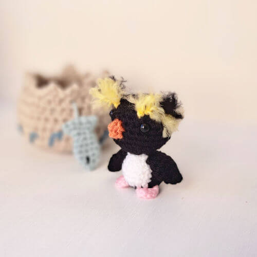 A rockhopper penguin crochet art figure standing next to his embroidered nest and fish