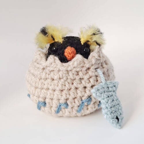 A rockhopper penguin crochet art figure peeping out of his embroidered nest with a little crochet fish hanging from the nest